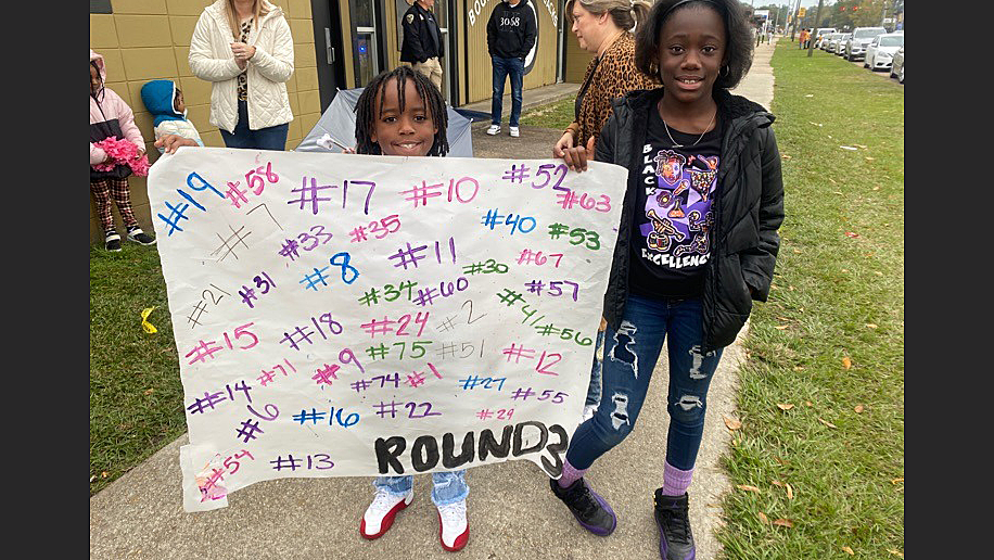 Two young supporters at a Bogalusa High School football team send-off event, proudly displaying a handmade sign with the jersey numbers of the players, symbolizing community support and school spirit ahead of a crucial playoff game.